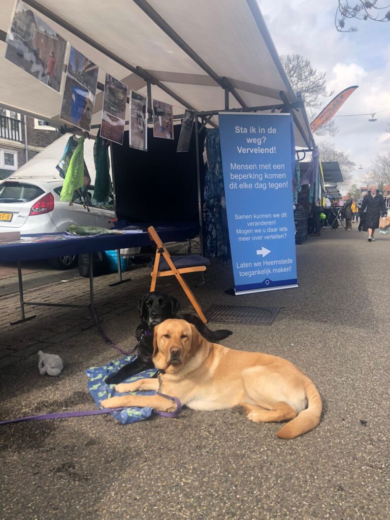Marktkraam met foto's, banner met tekst en geleidehond op de voorgrond